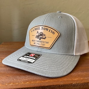May include: Gray and white trucker hat with a brown leather patch that says "Camel Towing" with a cartoon tow truck and the text "When it's wedged in tight we'll pull it out".