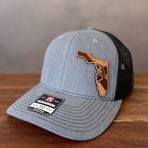May include: A gray and black trucker hat with a leather patch in the shape of Florida. The patch features a detailed design and the hat is adjustable. The Richardson brand logo is visible on the front.
