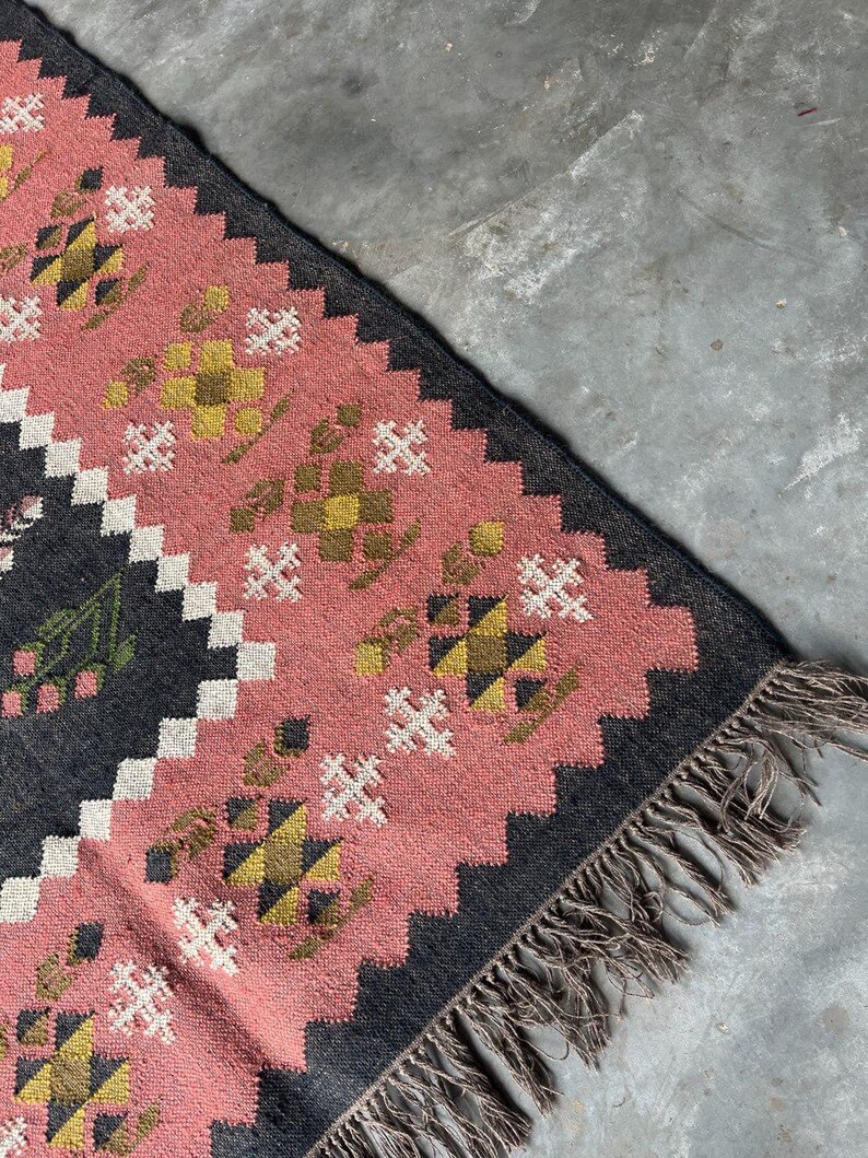 Op de afbeelding: Een rechthoekig tapijt met een roze en zwart geometrisch patroon. Het tapijt heeft een herhalend ontwerp van bloem- en ruitvormen in roze, geel en wit. Het tapijt heeft een gefranjerde rand en is gemaakt van geweven materiaal.