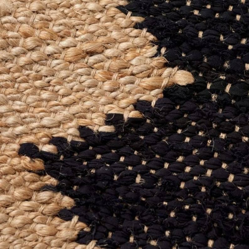 May include: Close-up of a woven rug with a geometric pattern. The rug features a combination of natural beige and black fibers, creating a textured surface. The design includes diagonal lines and shapes.