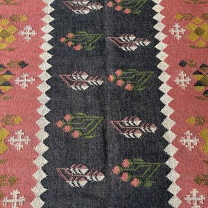 Op de afbeelding: Een rechthoekig tapijt met een centraal donkergrijs paneel met bloemmotieven in roze, groen en wit. Het tapijt heeft een roze rand met geometrische ontwerpen en een witte zigzagrand. Decoratief textiel.