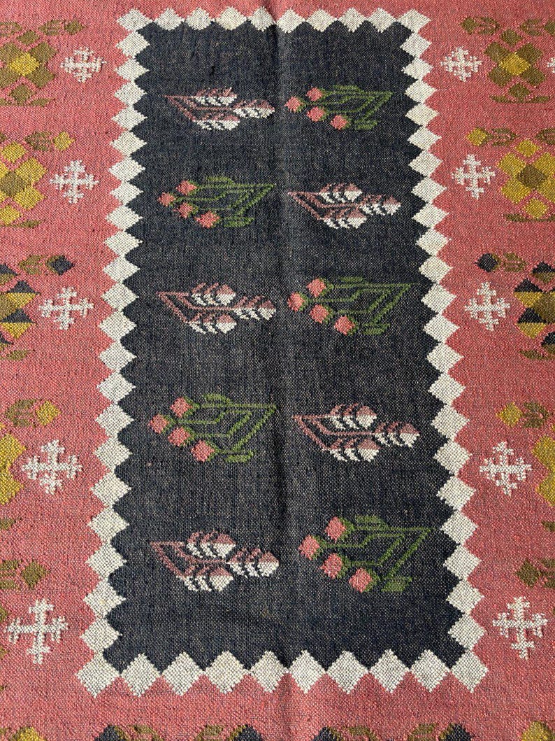 Op de afbeelding: Een rechthoekig tapijt met een centraal donkergrijs paneel met bloemmotieven in roze, groen en wit. Het tapijt heeft een roze rand met geometrische ontwerpen en een witte zigzagrand. Decoratief textiel.