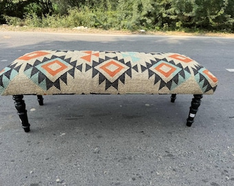 Puede incluir: Banqueta tapizada con un patrón geométrico en naranja, negro y turquesa sobre un fondo beige. La banqueta tiene cuatro patas torneadas negras. Una pieza decorativa para una sala de estar o entrada.