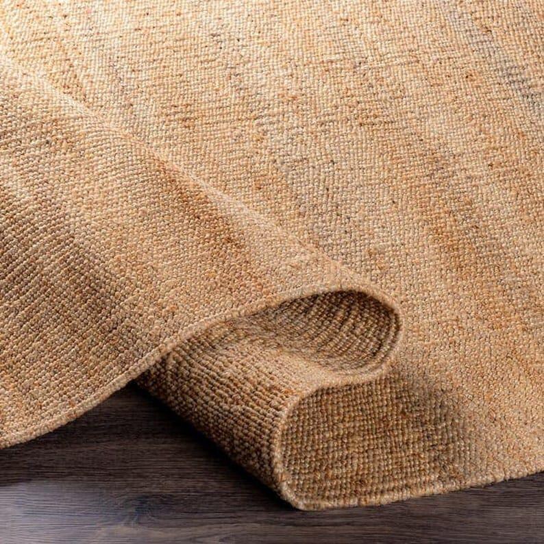 Op de afbeelding: Een close-up van een vloerkleed van natuurlijke vezels met een geweven textuur. Het vloerkleed is lichtbruin van kleur en heeft een licht ruw oppervlak.