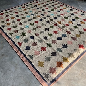 May include: A large, rectangular rug with a cream-coloured background and a pattern of colourful, geometric shapes. The rug has a pink and blue border.