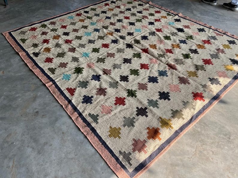 May include: A large, rectangular rug with a cream-coloured background and a pattern of colourful, geometric shapes. The rug has a pink and blue border.