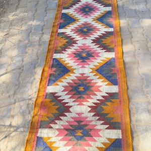 May include: A long, rectangular rug with a geometric pattern in shades of pink, blue, yellow, and grey. The rug has a fringe border.