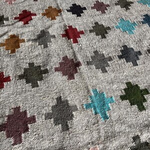 May include: A close-up of a woven rug with a geometric pattern. The rug features a variety of colours, including grey, blue, green, red, and brown. The pattern is made up of repeating crosses.