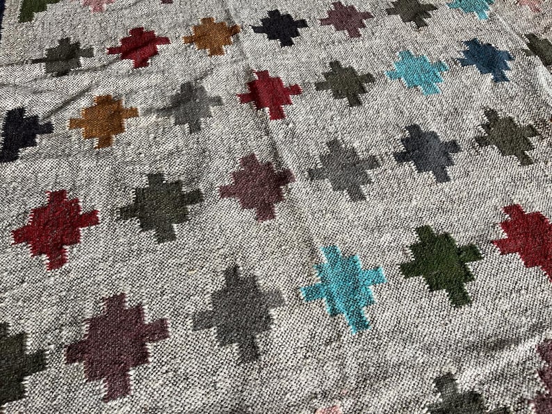 May include: A close-up of a woven rug with a geometric pattern. The rug features a variety of colours, including grey, blue, green, red, and brown. The pattern is made up of repeating crosses.