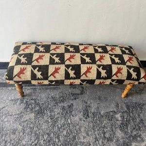 Può includere: Panca rettangolare con piano fantasia e gambe in legno. La parte superiore presenta un motivo ripetuto di forme rosse e bianche su uno sfondo a scacchi nero e beige. Le gambe sono marroni chiare.