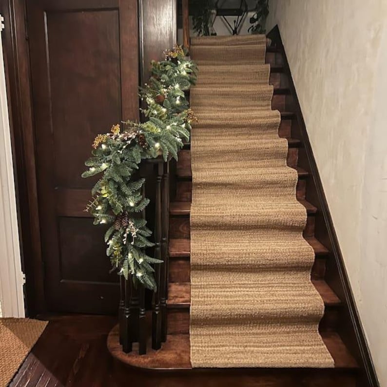 Op de afbeelding: Een houten trap met een bruine sisal loper. Een groene krans met witte lampjes is langs de leuning gedrapeerd.