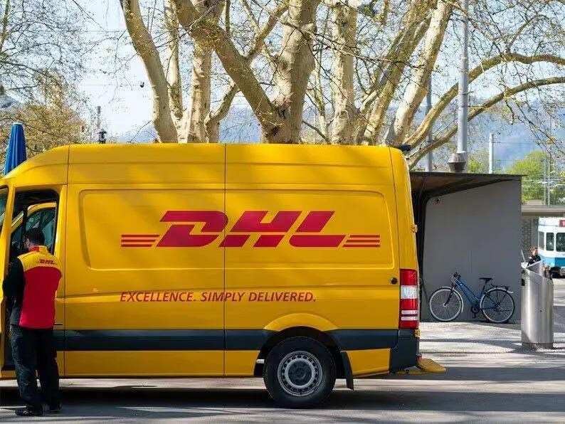 May include: A yellow DHL delivery van with the DHL logo in red. The van has the text "EXCELLENCE. SIMPLY DELIVERED." on the side. A person in a red and yellow vest stands near the van. A bicycle is parked nearby.