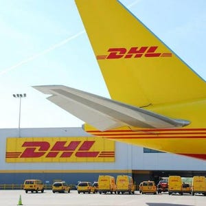 May include: A yellow DHL airplane tail with the DHL logo in red. The image also shows a building with the DHL logo and several yellow DHL vans parked in front of it, under a blue sky.