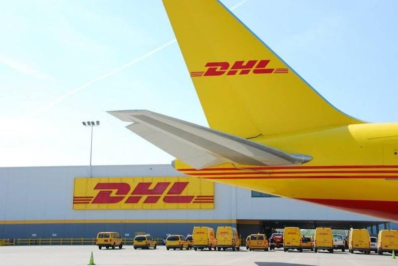 May include: A yellow DHL airplane tail with the DHL logo in red. The image also shows a building with the DHL logo and several yellow DHL vans parked in front of it, under a blue sky.
