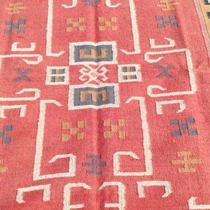 May include: A vintage red and beige rug with a geometric pattern. The rug has a central design with a cross shape and a border with a repeating pattern of leaves.