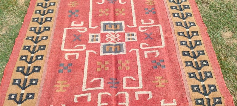 May include: A vintage red and beige rug with a geometric pattern. The rug has a central design with a cross shape and a border with a repeating pattern of leaves.