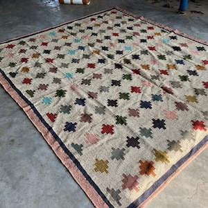 May include: A beige rug with a geometric pattern of colourful squares. The rug has a dark blue border with a pink trim.