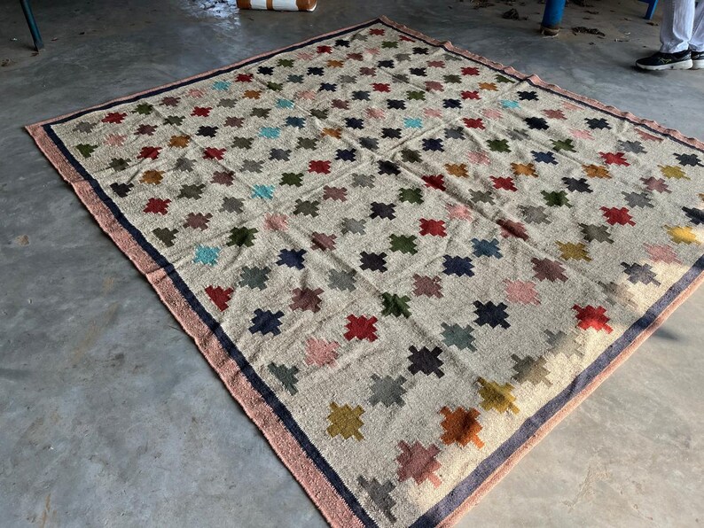 May include: A beige rug with a geometric pattern of colourful squares. The rug has a dark blue border with a pink trim.
