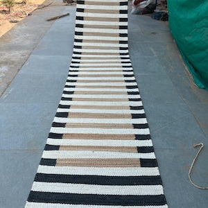 Puede incluir: Una alfombra de yute rayada en blanco y negro con un borde marrón natural. La alfombra está tejida con fibras gruesas y duraderas y tiene un diseño simple y moderno.