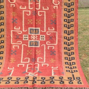 May include: A red, brown, and black patterned rug with a geometric design. The rug is made of natural fibers and has a fringed edge.