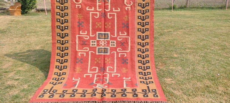 May include: A red, brown, and black patterned rug with a geometric design. The rug is made of natural fibers and has a fringed edge.