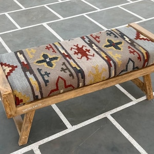 May include: Wooden bench with a rectangular seat featuring a patterned textile in gray, red, yellow, and black. The bench has a natural wood frame and angled legs, suitable for indoor or outdoor use.