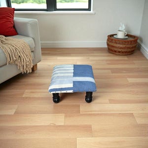 Puede incluir: Un pequeño taburete cuadrado con una parte superior estampada en azul y blanco. El diseño de la tela presenta rayas y formas geométricas. El taburete tiene cuatro patas cortas, negras y redondeadas. Una pieza decorativa para una sala de estar o dormitorio.