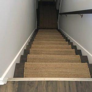 Op de afbeelding: Natuurlijke vezel traptreden in een lichtbruine kleur, die een set houten trappen met een donkerbruine afwerking bedekken. De treden zijn geweven met een visgraatpatroon en hebben een licht getextureerd oppervlak.
