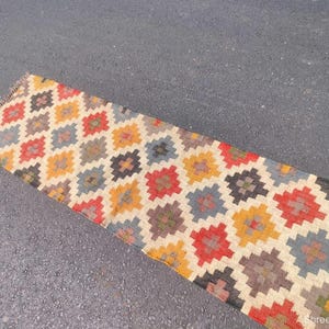 May include: A rectangular, patterned rug with a repeating geometric design. The rug features a beige background with colorful diamond-shaped patterns in red, yellow, blue, and brown. The rug is placed on a gray surface.