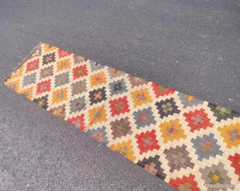 May include: A rectangular, patterned rug with a repeating geometric design. The rug features a beige background with colorful diamond-shaped patterns in red, yellow, blue, and brown. The rug is placed on a gray surface.