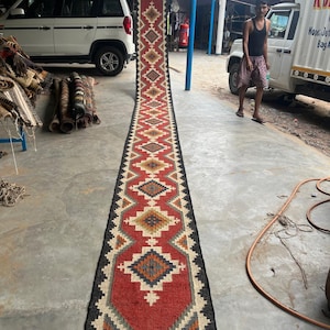 May include: A long, narrow, red, black, white, and orange rug with a geometric pattern. The rug is hanging on a wall in a shop. The text "shree Balaji Rugs" is visible at the bottom of the rug.