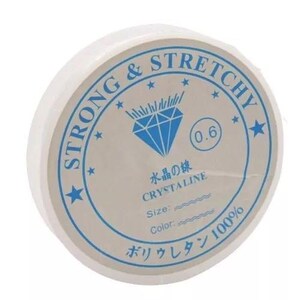May include: A spool of white, stretchy beading cord. The label is white with blue text and a diamond graphic. Text on the label includes "STRONG & STRETCHY", "CRYSTALINE", and Japanese characters. The cord is 0.6 mm in diameter.