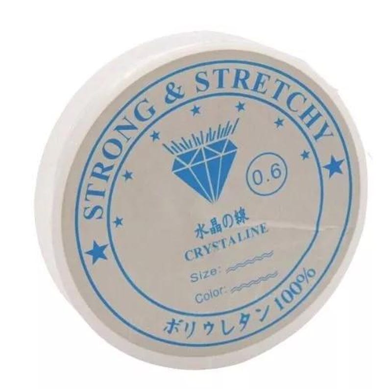 May include: A spool of white, stretchy beading cord. The label is white with blue text and a diamond graphic. Text on the label includes "STRONG & STRETCHY", "CRYSTALINE", and Japanese characters. The cord is 0.6 mm in diameter.
