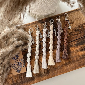 May include: Six handmade macrame keyrings with different coloured tassels and charms. The keyrings are made with a white, cream, light brown, grey, and brown cord. The charms include a clover, a Star of David, and a small bell.