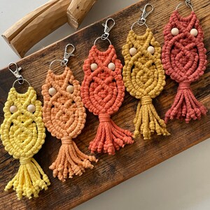 May include: Five colourful macrame owl keychains with wooden beads for eyes. The keychains are arranged in a gradient from yellow to orange to red.