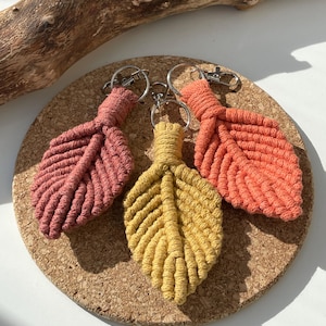 May include: Three macrame leaf keyrings in coral, peach, and yellow. The keyrings are made with a knotted cord and have a silver metal ring.