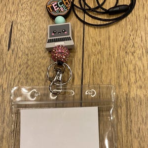 Puede incluir: Un cord&oacute;n negro con una cuenta de silicona en forma de manzana con el texto TECH VIBES, una cuenta redonda azul claro, una cuenta gris con forma de port&aacute;til con una cara, una cuenta de estr&aacute;s rosa, un cierre de metal plateado y un portatarjetas de pl&aacute;stico transparente.