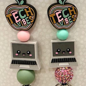 May include: Two lanyards featuring black cords, silver metal lobster clasps, and silicone charms. Each charm set includes an apple shape with the text TECH VIBES, a round bead, and a grey laptop with a smiling face. One has a pink bead, one a mint bead, and one a pink rhinestone bead.