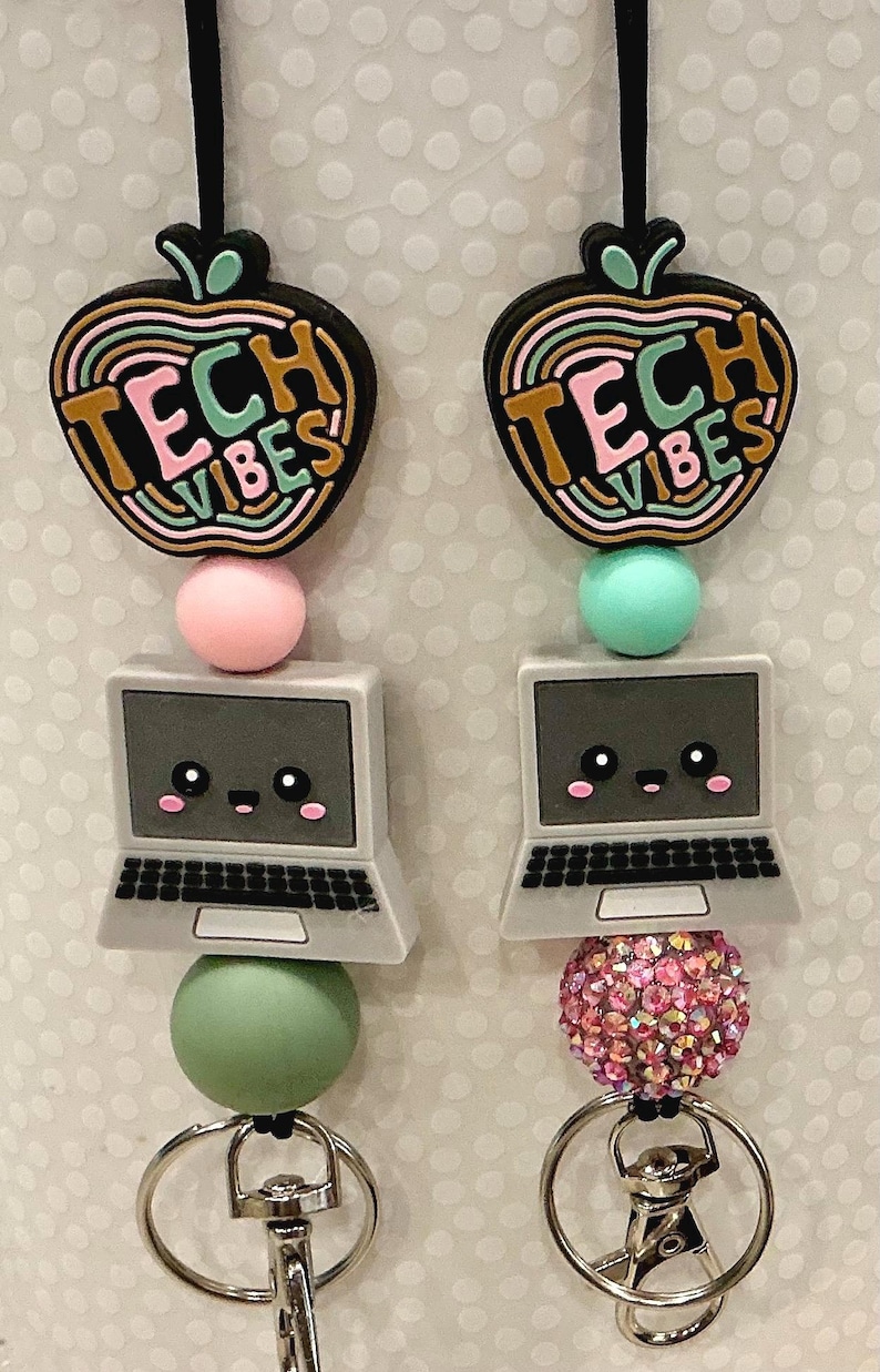 Puede incluir: Dos cordones con cierres de mosquet&oacute;n de metal plateado y dijes de silicona. Cada conjunto incluye una forma de manzana con el texto TECH VIBES, una cuenta redonda y una computadora port&aacute;til gris con una cara sonriente. Las cuentas son rosa, menta y de pedrer&iacute;a rosa.
