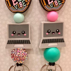 以下が含まれることがあります： 黒い紐、カラフルなリンゴのチャーム（「Tech Vibes」の文字入り）、笑顔のノートパソコンのチャーム、キラキラ光るビーズが付いた、テクノロジーをテーマにしたキーホルダーが2つ。1つのキーホルダーにはピンクのビーズとターコイズブルーのビーズが付いており、もう1つのキーホルダーにはターコイズブルーのビーズとピンクのキラキラ光るビーズが付いています。
