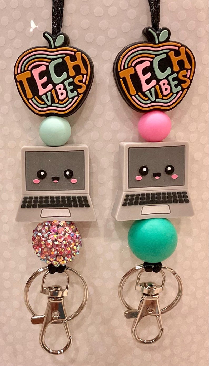 Puede incluir: Dos llaveros con un dije negro en forma de manzana con el texto TECH VIBES en colores arco&iacute;ris, un dije de computadora gris con cara sonriente y cuentas de silicona coloridas. Uno tiene una cuenta de diamantes de imitaci&oacute;n y ambos tienen broches plateados.