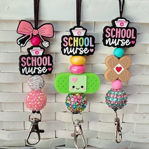 Puede incluir: Tres cordones con cuentas con el texto "SCHOOL nurse". Cada cordón presenta un diseño diferente, que incluye un lazo rosa, una venda y una venda en forma de cruz. Los cordones tienen una variedad de cuentas coloridas y un cierre plateado.