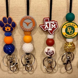 Puede incluir: Un conjunto de diez cordones coloridos con diferentes logotipos universitarios y cuentas. Cada cordón tiene un cierre de seguridad negro y un llavero plateado. Los logotipos incluyen: Tennessee, Auburn, Texas Longhorns, Texas A&M, Baylor University, Ole Miss y Carolina del Norte.