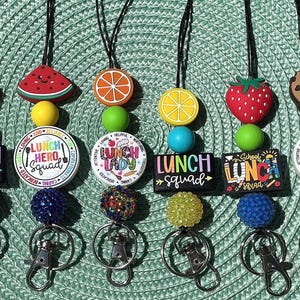 Puede incluir: Seis cordones con cuentas de silicona de colores, dijes con forma de frutas como uvas, sandía, naranja, limón, fresa y una galleta, junto con colgantes redondos y rectangulares impresos con el texto Lunch Hero Squad y Lunch Lady.