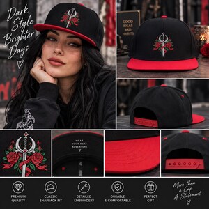 May include: A black snapback cap with a red brim featuring an embroidered silver sword entwined with red roses and green leaves. The interior brim displays the text "WEAR YOUR NEXT ADVENTURE" with a heart symbol. The back includes a red plastic snap closure.