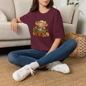 May include: A maroon short-sleeved t-shirt featuring a graphic of an elderly person in a straw hat watering several potted Venus flytrap plants with a blue watering can, accompanied by a small snail and a bag labeled Plant Food.