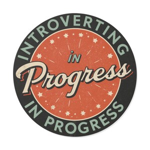 May include: Round sticker with the text "INTROVERTING IN PROGRESS" in a vintage style. The design features a red center with the words "in Progress" in a stylized font, surrounded by a black border and small star accents.