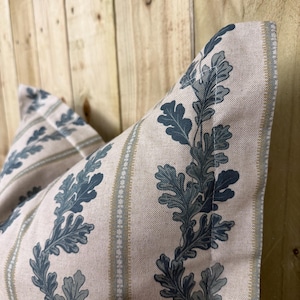 May include: Close-up of two decorative cushions. The cushions feature a beige background with vertical stripes and a repeating pattern of blue-green oak leaves. The cushions have a soft, textured appearance, suggesting a comfortable design.