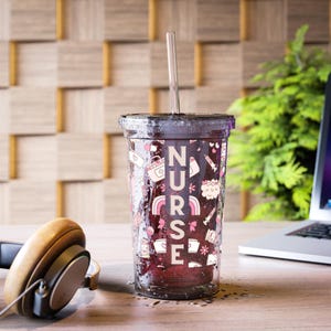 May include: A clear tumbler with a black lid and a silver straw. The tumbler is decorated with the word "NURSE" in pink letters and various medical-themed illustrations. A pair of brown headphones and a laptop are in the background.