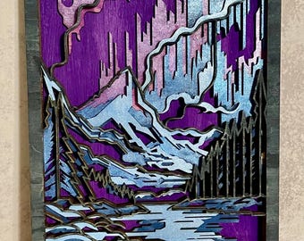 May include: A layered wooden artwork showing a mountain landscape with an aurora borealis. The piece uses shades of purple, blue, and silver, with a dark grey frame. The design includes mountains, trees, and a body of water.